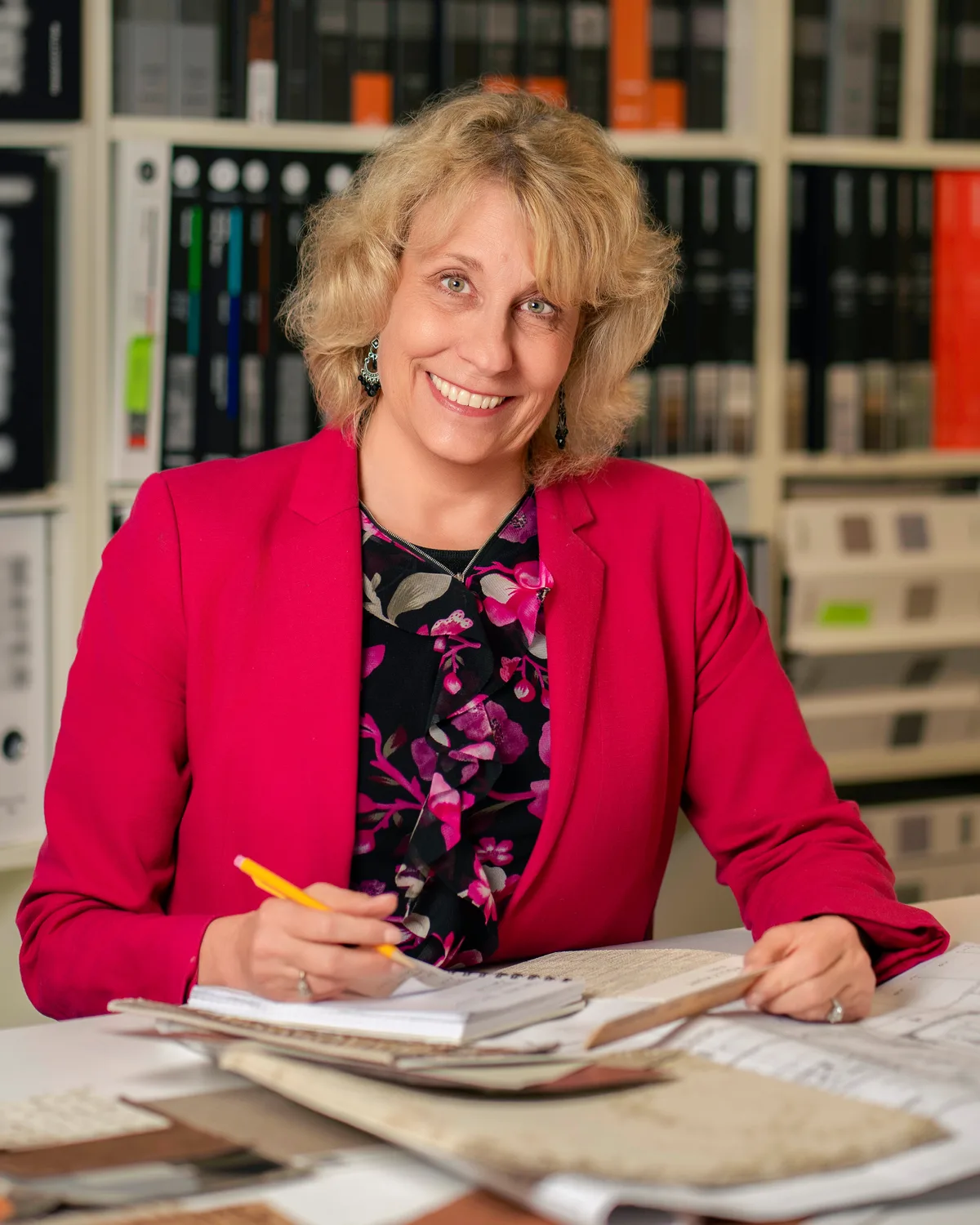 A designer at her worktable with fabric samples and a pencil, shelves of sample binders behind her.
