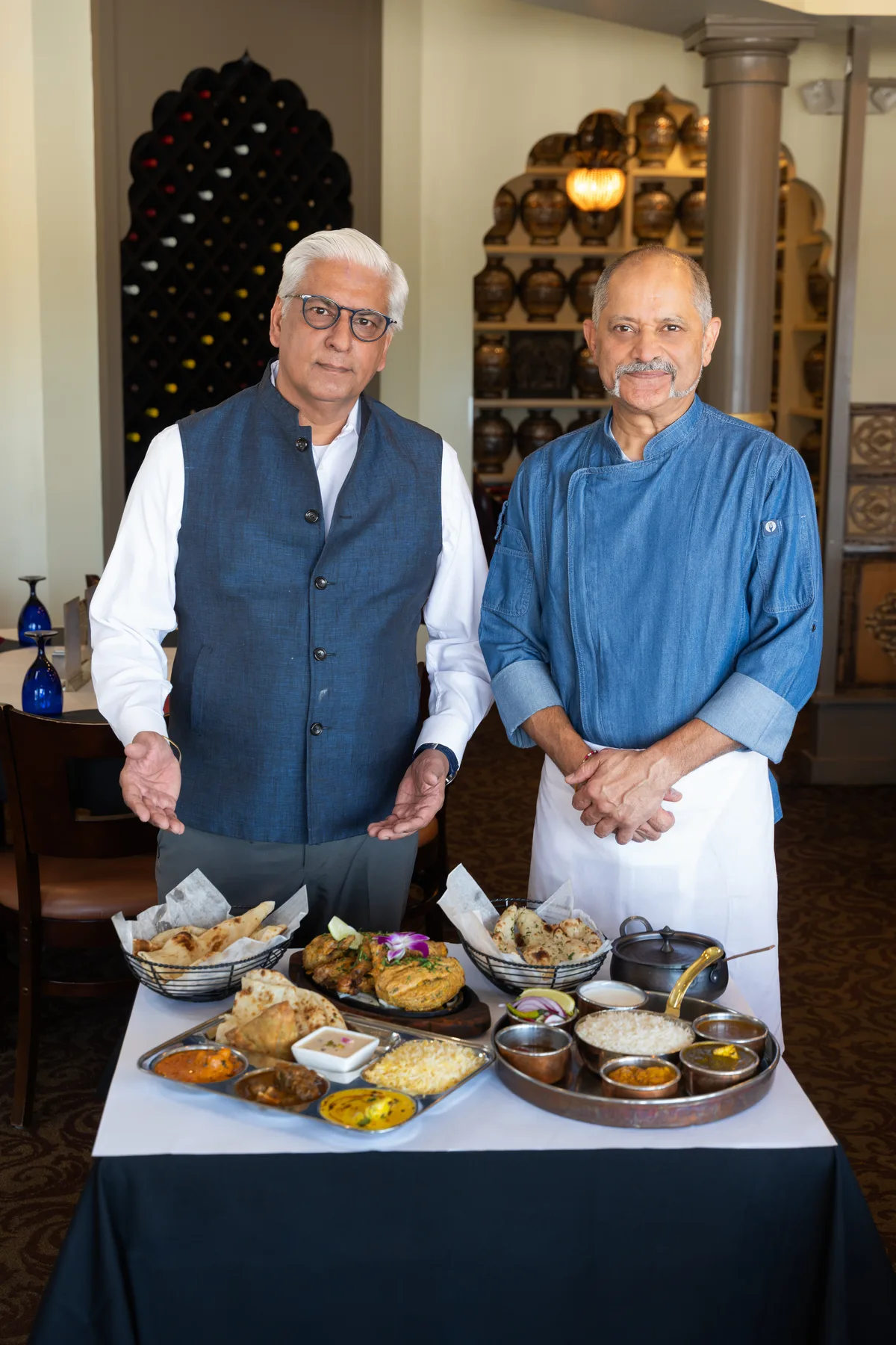 Two restaurant owners behind a table set with thali platters, naan and curries in their dining room.